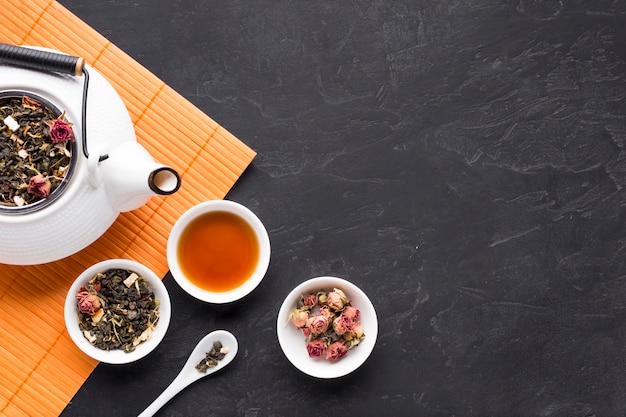 Dry roses and tea herb with teapot on orange placemat over black stone backdrop