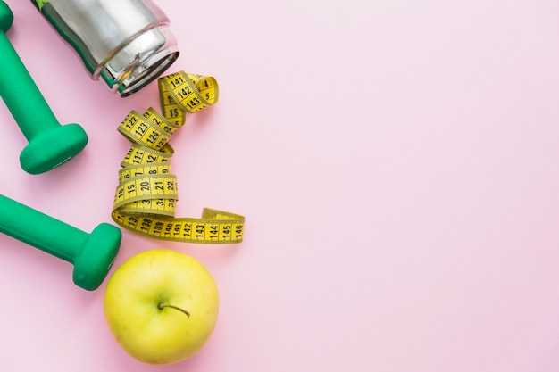 Free Photo dumbbells; water bottle; measuring tape and apple on pink background