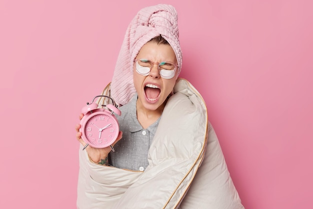 Free photo early awakening concept. sleepy young european woman yawns and keeps mouth widely opened holds alarm clock wrapped in blanket wears bath towel on head after taking shower isolated over pink background