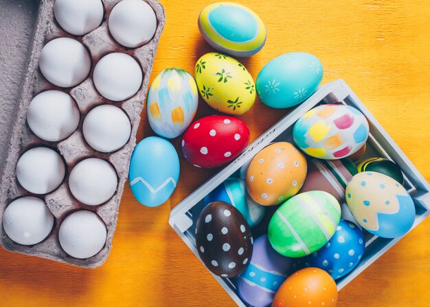 Easter eggs in wooden box and egg carton top view on yellow wooden background