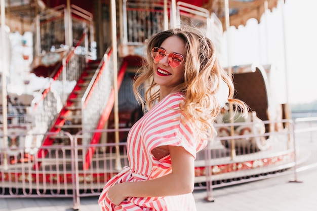 Easy-going woman in pink sunglasses spending weekend in amusement park. Graceful white girl having fun in summer vacation.