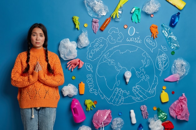 Free photo ecology and land conservation concept. sad asian woman stands in praying pose, surrounded with plastic waste, begs for help in cleaning earth, dressed casually