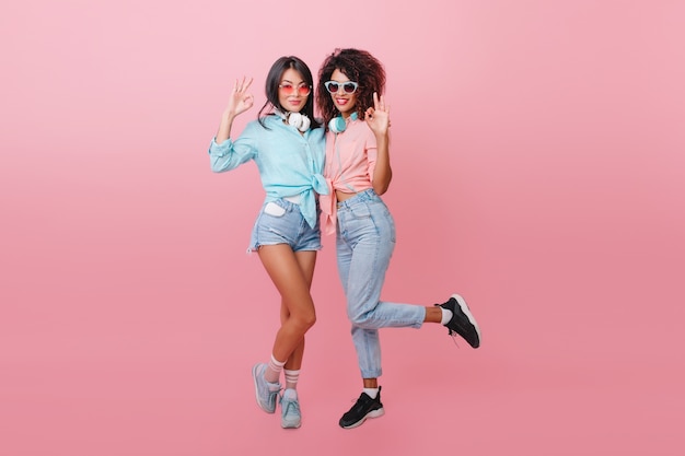 ecstatic black woman in sport shoes jumping near european slim friend. Elegant asian girl enjoying free time with mulatto lady in cotton shirt.