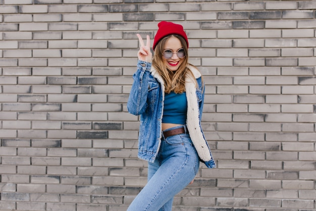 Ecstatic blonde girl in red hat standing in confident pose beside brick wall. Outdoor shot of cute caucasian woman in jeans and denim jacket having fun on the street.