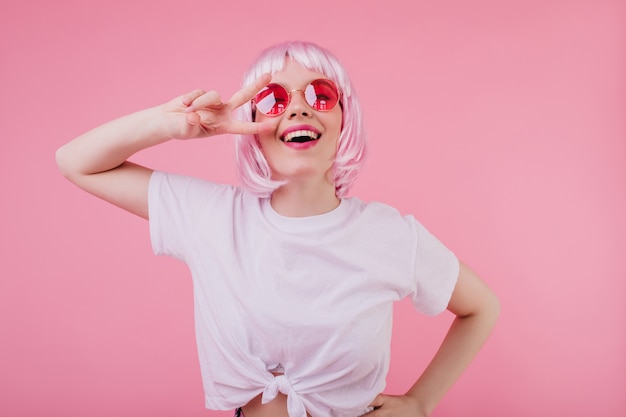 Ecstatic caucasian girl in trendy white t-shirt posing with peace sign and laughing. Indoor photo of dreamy european woman in shiny peruke and sunglasses