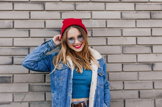 Ecstatic caucasian model with light-brown hair posing with smile beside brick wall. Outdoor portrait of carefree young woman wears trendy denim jacket.