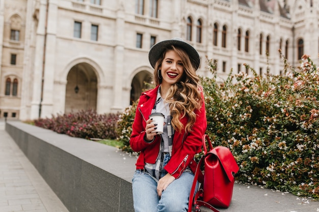 Ecstatic curly woman in red jacket drinking coffee in front of beautiful old building