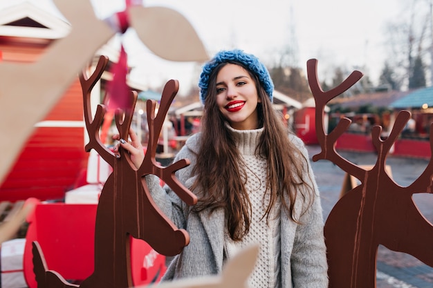 Free photo ecstatic dark-haired female model enjoying christmas in themed amusement park. outdoor portrait of glad girl in knitted blue hat posing near holiday decoration in winter.