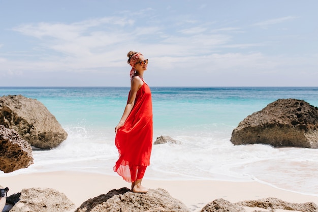 Ecstatic girl with pretty smile standing on big stone with ocean. Outdoor full-length photo of cheerful female tourist chilling at wild beach.