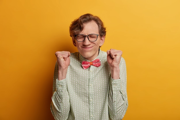 Free photo ecstatic male nerd raises clenched fists, believes in success, closes eyes and awaits announcement of results, participates in contetst, hopes to win, expresses impatience, dressed formally.