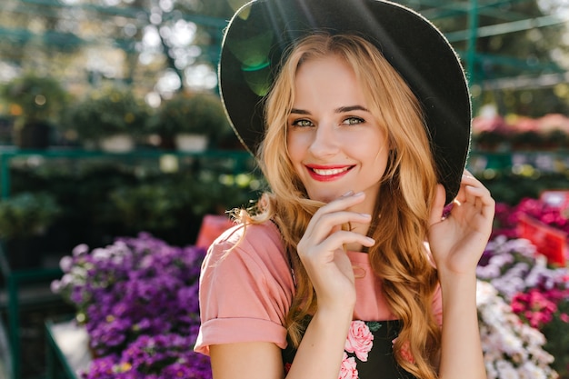 Ecstatic young woman with light eyes posing near flowers. Cute european woman in hat chilling on orangery.