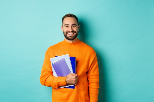 Education. Smiling bearded man holding notebooks and smiling, going on courses, standing over light turquoise wall