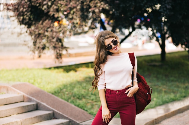 Effective girl with a long curly hair in vinous pants is posing  on street in the city.