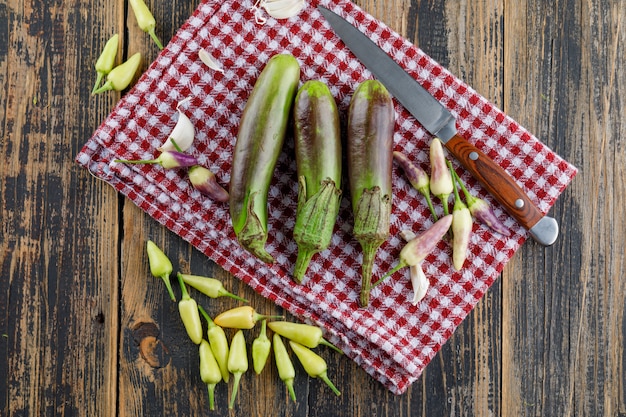 Eggplants with garlic, peppers, knife on wooden and kitchen towel, flat lay.