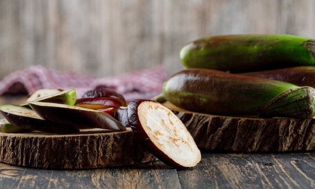 Free photo eggplants with slices, wood pieces side view on wooden and kitchen towel