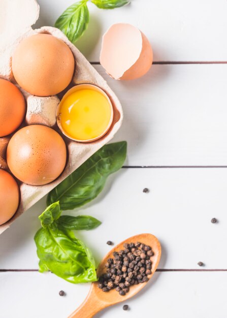 Eggs in carton with basil leaf and peppercorn on wooden plank