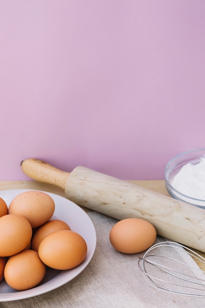 Eggs on plate; rolling pin; whisk and flour on desk against pink background