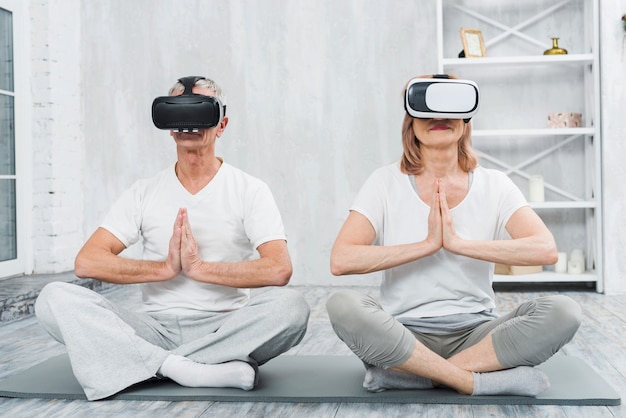 Elder couple wearing virtual reality headset in sitting on mat with praying hands gesture