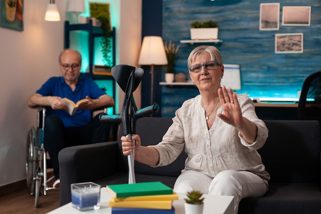 Elder husband and wife with crutches and wheelchair sitting on couch at home. Caucasian old man reading book and woman waving hand at camera on video call technology device gadget