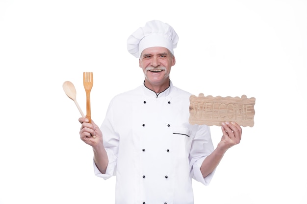 Elderly chief man in cook uniform smiling while holding welcome plate, spoon and fork isolated over white wall