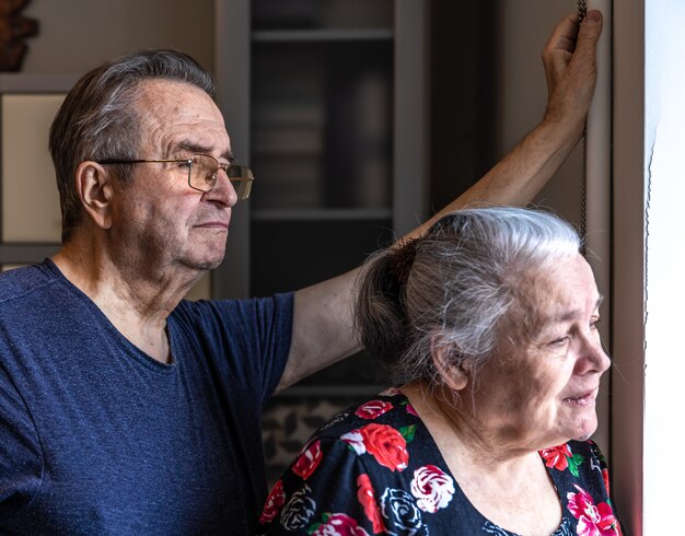 An elderly couple stands at the window and looks out at someone.