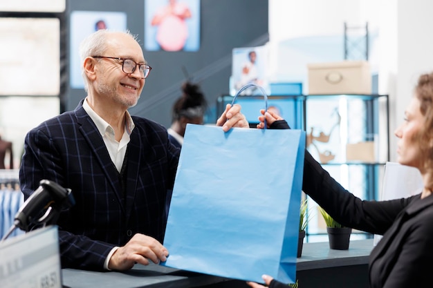 Elderly customer taking shopping bag from store employee, standing at counter desk paying for purchase in fashion boutique. Senior shopper buying stylish clothes making electronic transaction at mall