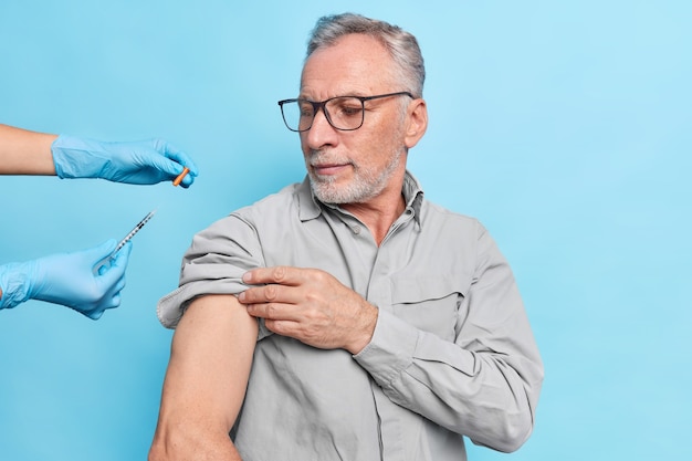 Free Photo elderly man gets vaccination against coronavirus looks attentively at syringe with vaccine wears eyeglasses