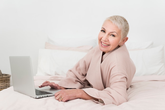 Elderly woman in bathrobe smiling and posing with laptop in bed