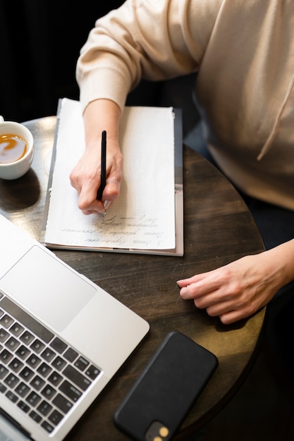 Elderly woman drinking coffee at a cafe while working on her laptop and writing on her notebook