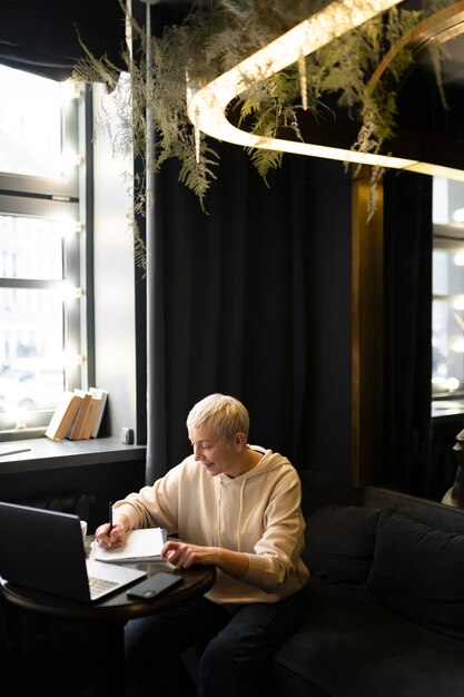 Elderly woman drinking coffee at a cafe while working on her laptop and writing on her notebook