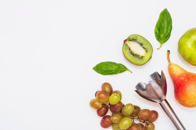 Free photo an electric hand blender with kiwi; apple; pear; grapes and basil leaves on white background