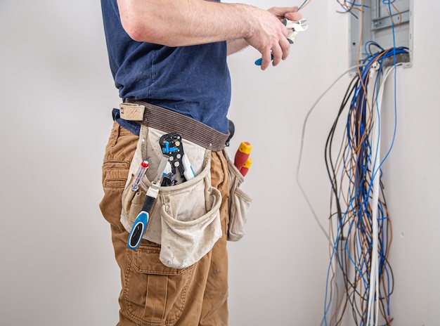 Electrician builder at work, examines the cable connection in the electrical line in the fuselage of an industrial switchboard. Professional in overalls with an electrician's tool.