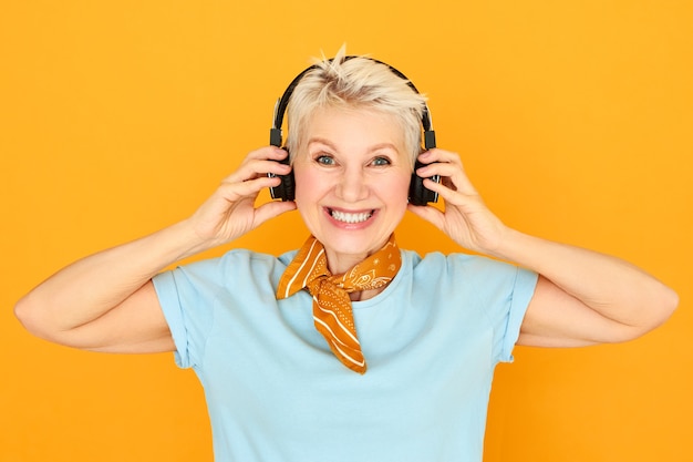 Free photo electronic devices, entertainment, retirement and age concept. charming happy blonde woman pensioner wearing black wireless headphones, enjoying nice high resolution audio sound, listening to music