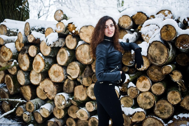 Elegance curly girl in leather jacket at snowy forest park at winter against stumps