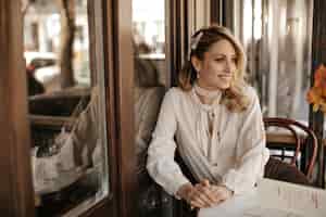 Free photo elegant beautiful blonde woman in white stylish blouse, pearl jewelry smiles widely, looks away and sits by little table in street cafe