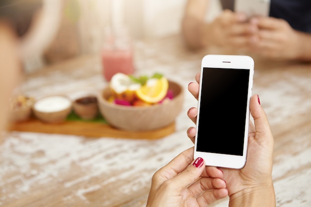 Elegant female hands with neat red nails using white mobile phone, viewing newsfeed via her account on social networks