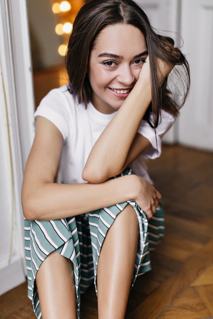 Elegant girl posing in pajamas in good weekend morning. Brunette caucasian woman sitting on the floor with beautiful smile.