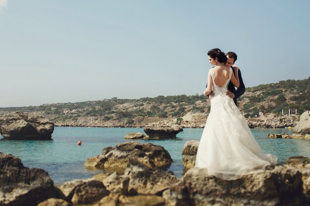 Elegant smiling young bride and groom posing on the rocks on the beach