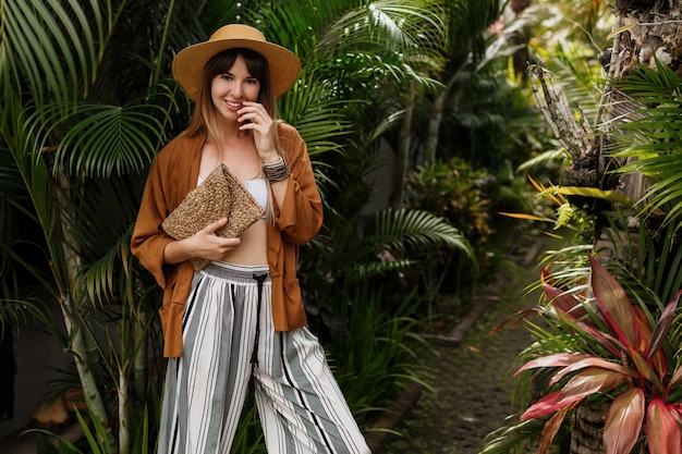 Free photo elegant stylish girl in white top and straw hat posing on palm leaves in bali.