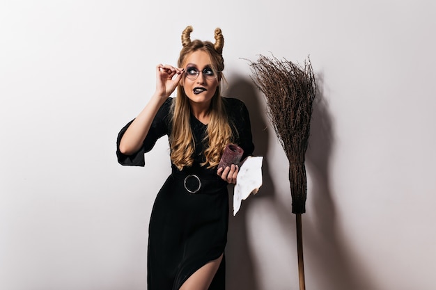 Free Photo elegant wizard in glasses funny posing on white wall. fair-haired witch standing near broom.