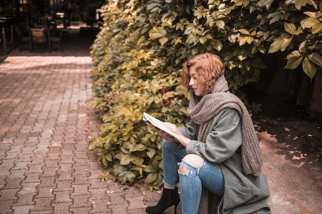 Free photo elegant woman sitting on board and attentively reading book near plant