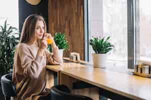 Free photo elegant young positive woman holding cup of drink at bar counter near window in cafe