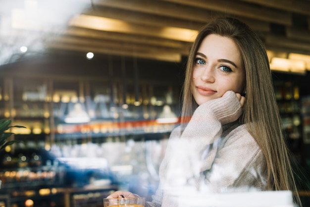 Elegant young positive woman with cup of drink in cafe through window