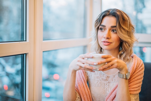 Free photo elegant young thoughtful woman with cup of drink near window in cafe