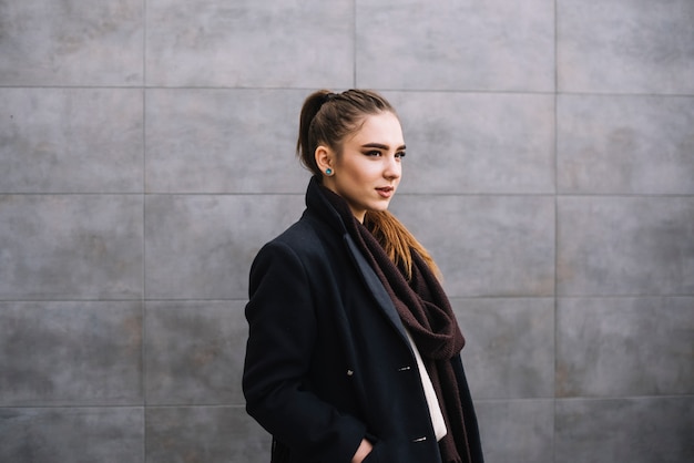 Free Photo elegant young woman in coat with scarf near grey wall