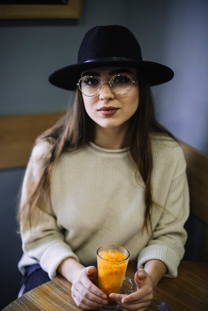 Elegant young woman in hat and eyeglasses with glass of drink