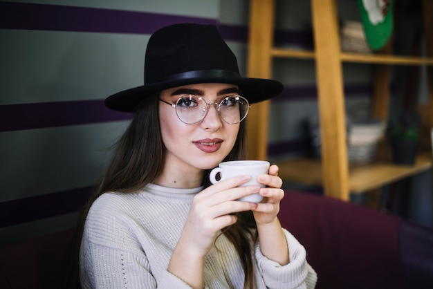 Elegant young woman in hat and eyeglasses with mug of drink