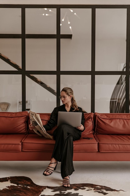 Free Photo elegant young woman in silk black jumpsuit and eyeglasses sits on brown sofa in living room reads