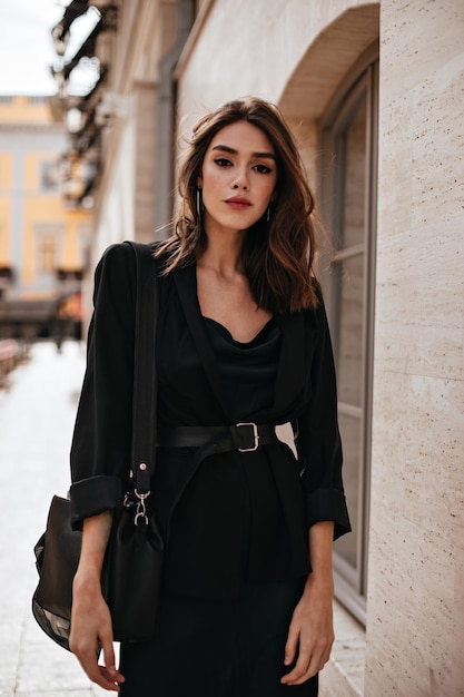 Free photo elegant young woman with brown wavy hair red lips eyeliner dark dress jacket and stylish black accessories walking on street at summer autumn and looking straight