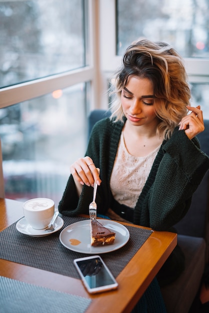 Free photo elegant young woman with dessert and cup of drink near smartphone at table